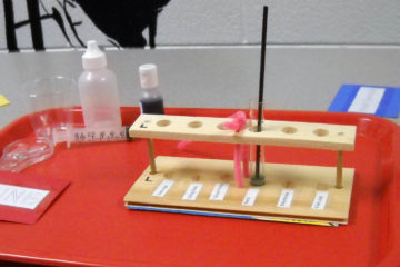 Photo of the testing to be given to each team of students for the Mystery Powders activity. Contained on the cafeteria tray are a test-tube rack, a test tube, a stirrer, a magnifying glass, a cup of water, a dropper bottle of vinegar, a dropper bottle of bromothymol blue, a syringe, and a set of chemical cards for requesting the powder samples and iodine